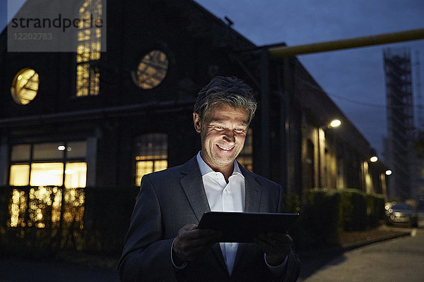 Lächelnder Geschäftsmann mit Tablette vor dem modernen Gebäude bei Nacht