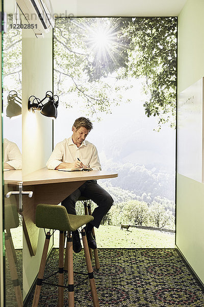 Geschäftsmann beim Schreiben in Buch am Tisch im modernen Büro mit Panoramafenster