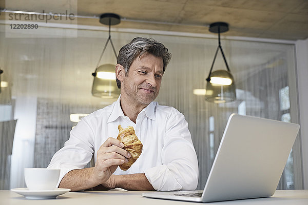 Geschäftsmann mit Laptop beim Croissantessen
