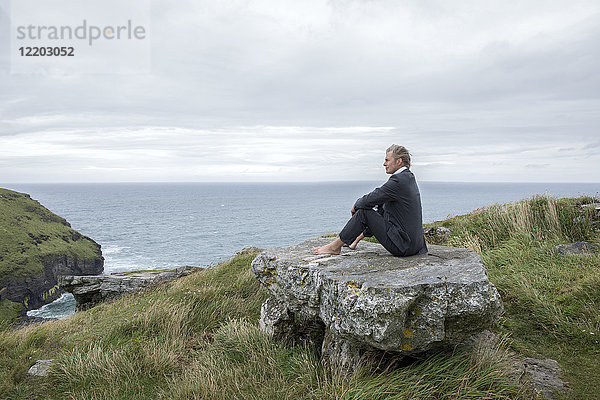 UK  Cornwall  Tintagel  Geschäftsmann auf einem Felsen an der Küste sitzend mit Blick auf die Aussicht