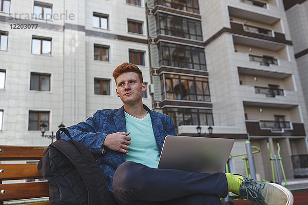 Rothaariger junger Mann sitzend auf Bank mit Laptop