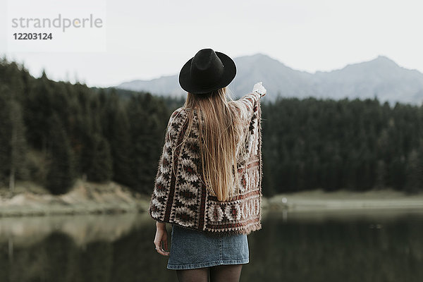 Rückansicht der modischen jungen Frau mit Hut und Poncho vor dem See