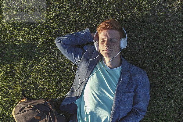Rothaariger junger Mann mit Kopfhörer auf Gras liegend