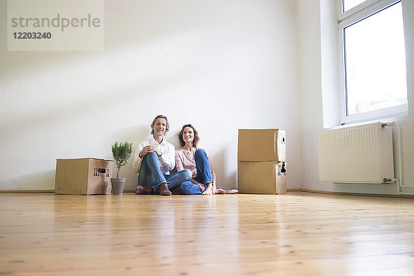 Lächelndes reifes Paar sitzt auf dem Boden in einem leeren Raum neben Pappkartons.