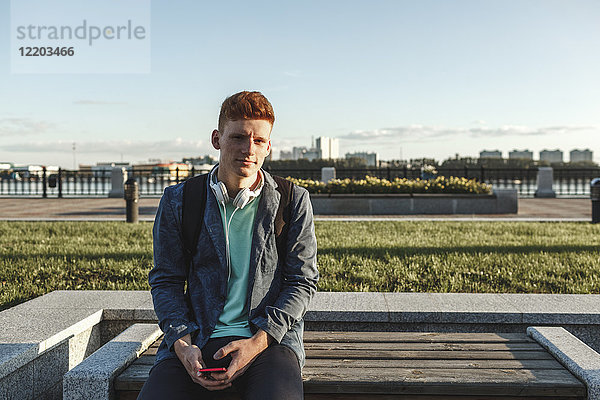 Porträt eines rothaarigen jungen Mannes auf der Bank mit Smartphone und Kopfhörer