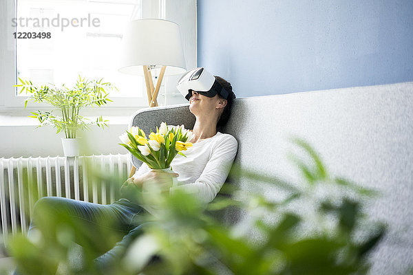 Lächelnde Frau sitzt auf der Couch und trägt eine VR-Brille mit Tulpen.