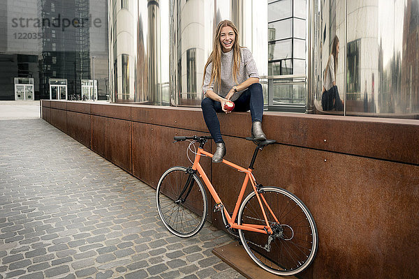 Fröhliche junge Frau mit Fahrrad bei einer Pause in der Stadt beim Apfelessen