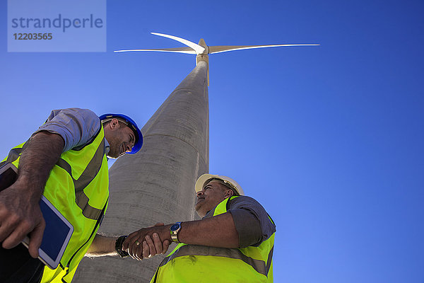 Niederwinkelansicht von Technikern  die vor der Windkraftanlage die Hand schütteln