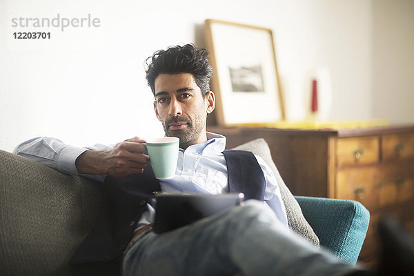 Porträt eines nachdenklichen Mannes mit Kaffeetasse und Tablette auf dem Sofa zu Hause