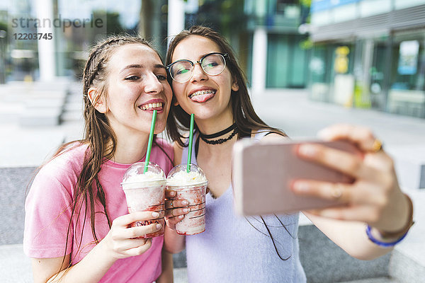 Zwei fröhliche Teenager-Mädchen trinken Milchshakes in der Stadt mit einem Selfie.