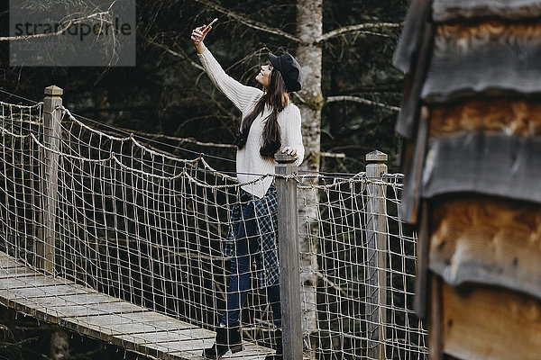Junge Frau auf einer Hängebrücke am Baumhaus im Wald mit einem Selfie