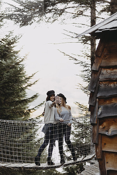 Zwei junge Frauen auf einer Hängebrücke bei einem Baumhaus im Wald mit einem Selfie