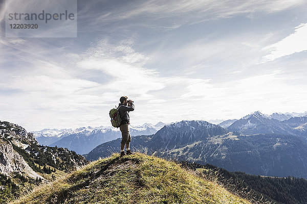 Österreich  Tirol  junger Mann steht in der Berglandschaft und schaut mit dem Fernglas in die Ferne.
