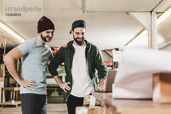 Zwei lächelnde junge Männer beim Blick auf den Laptop in der Werkstatt