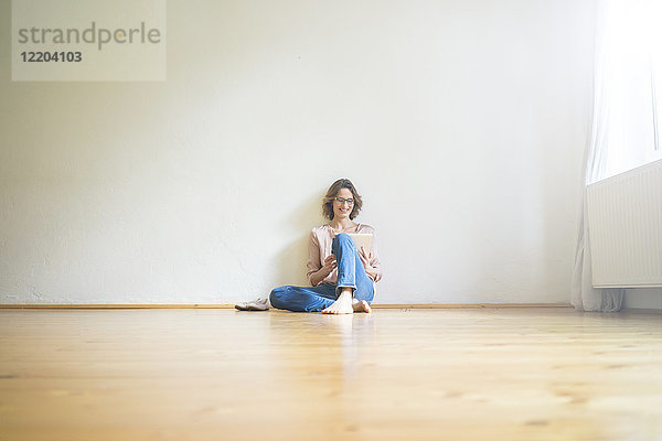 Lächelnde reife Frau sitzt auf dem Boden im leeren Raum mit Tablette