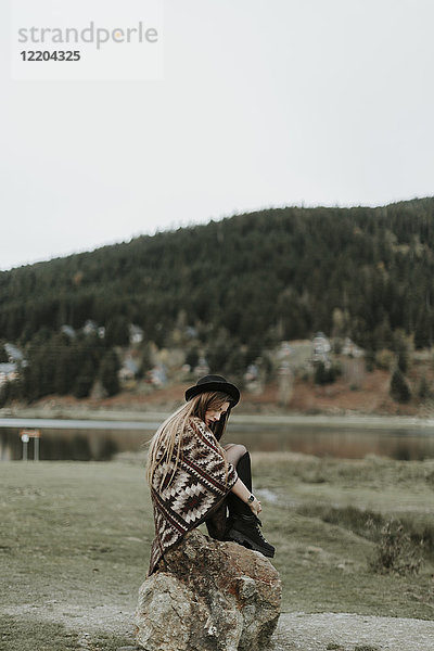 Modische junge Frau mit Hut und Poncho auf einem Felsen sitzend