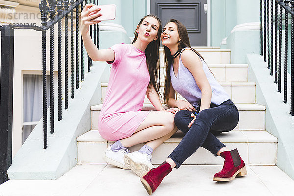 Zwei fröhliche Teenager-Mädchen sitzen auf einer Treppe in der Stadt und nehmen einen Selfie.