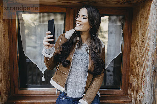 Fröhliche junge Frau am Fenster eines Holzhauses mit einem Selfie