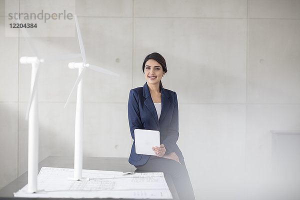Porträt einer lächelnden jungen Frau mit Bauplan  Windkraftanlagenmodellen und Tablette im Büro