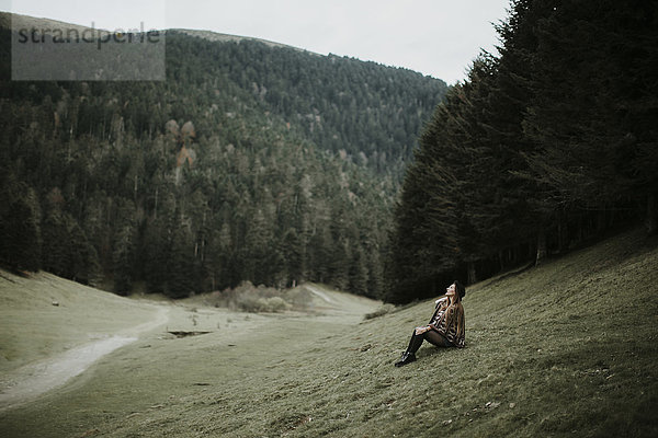 Junge Frau sitzt auf einer Wiese am Waldrand und schaut nach oben.