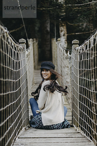Porträt einer lächelnden jungen Frau  die auf einer Hängebrücke sitzt.