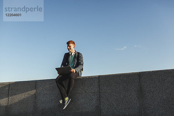 Lächelnder rothaariger junger Mann auf der Wand sitzend mit Laptop