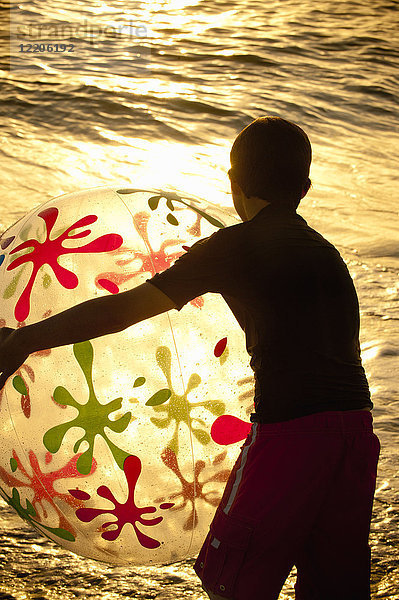 Silhouette eines kaukasischen Jungen  der bei Sonnenuntergang einen Strandball hält