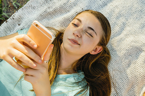 Verwirrtes kaukasisches Mädchen liegt auf einer Decke und schreibt eine SMS auf einem Handy