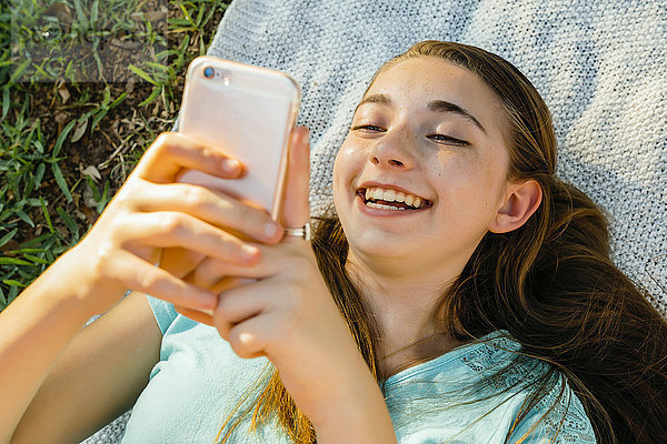 Lachendes kaukasisches Mädchen  das auf einer Decke liegt und eine SMS auf einem Handy schreibt
