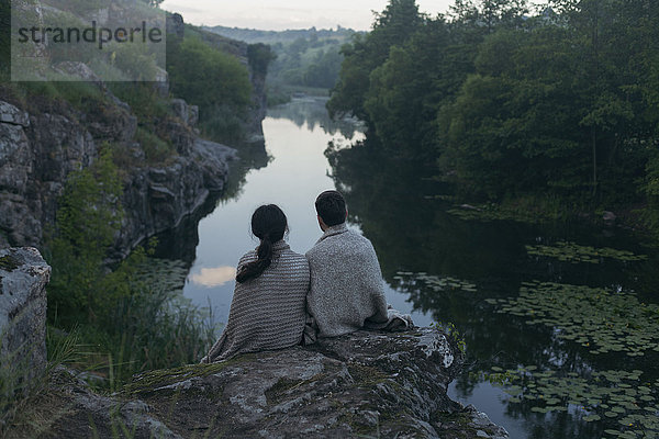 Kaukasisches Paar  eingewickelt in eine Decke  sitzt auf einem Felsen und bewundert den Fluss