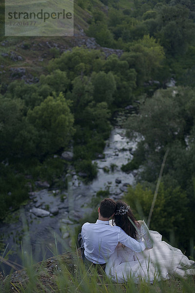 Kaukasische Braut und Bräutigam sitzen auf einem Hügel und bewundern den Fluss