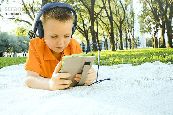 Ernster kaukasischer Junge  der auf einer Decke im Park liegt und einem digitalen Tablet zuhört