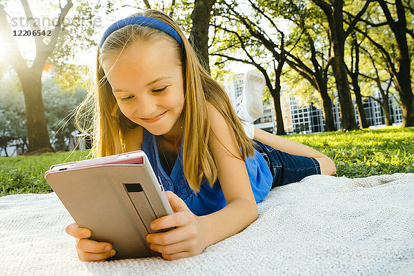 Lächelndes kaukasisches Mädchen  das auf einer Decke im Park liegt und ein digitales Tablet liest