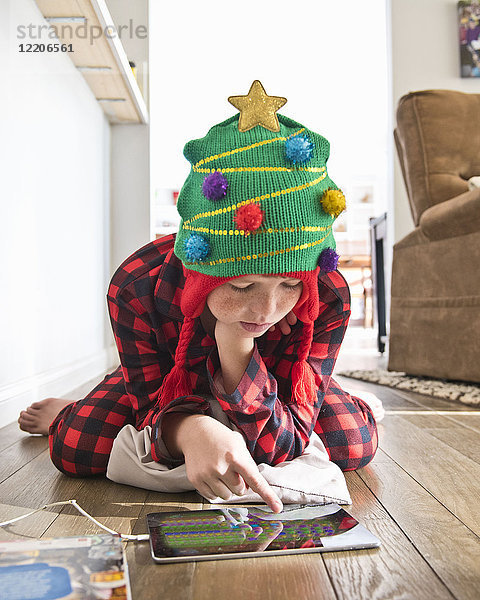 Kaukasischer Junge mit Weihnachtsbaum-Strumpfmütze  der ein digitales Tablet auf dem Boden benutzt