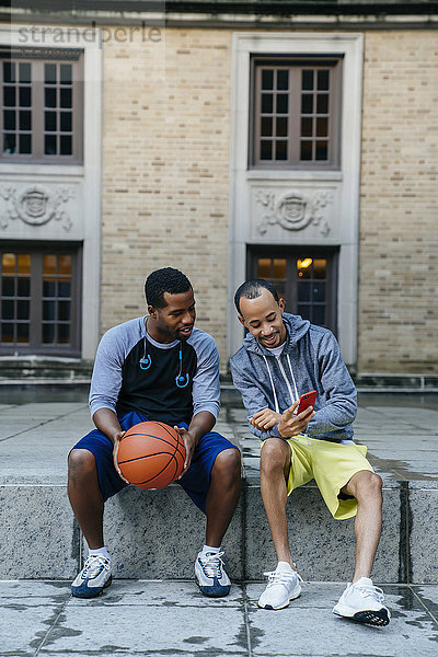 Schwarze Männer  die mit einem Basketball auf dem Beton sitzen und mit dem Handy SMS schreiben