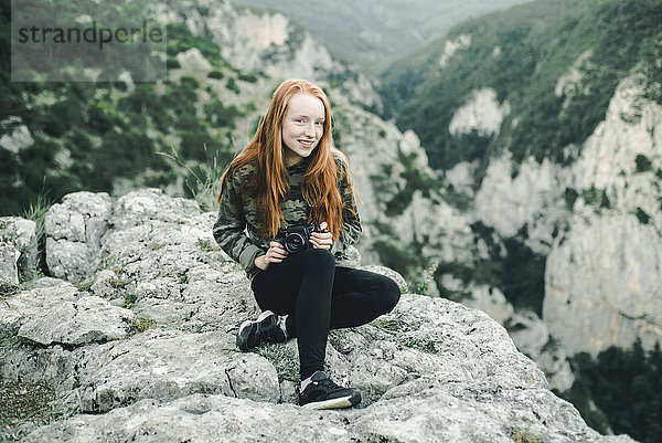 Porträt einer lächelnden kaukasischen Frau  die auf einem Felsen sitzt und eine Kamera hält
