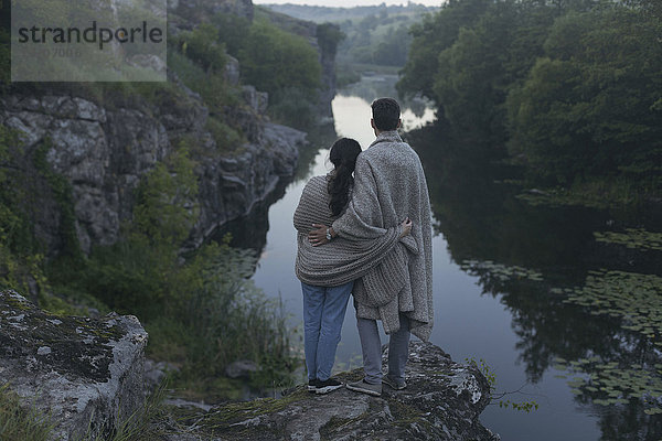 Caucasian couple wrapped in blanket standing on rock admiring river