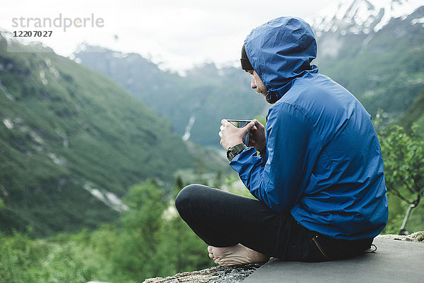 Kaukasischer Mann sitzt in einer Berglandschaft und trinkt Kaffee