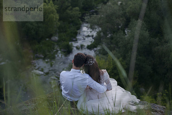 Caucasian bride and groom sitting on hill admiring river