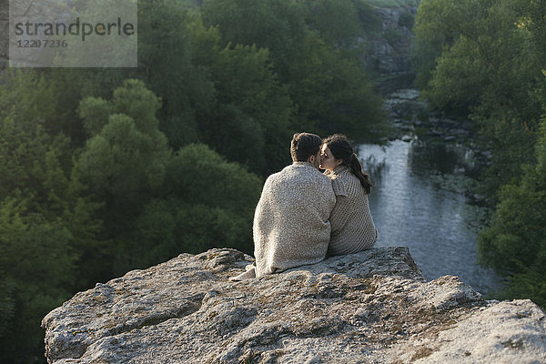 Caucasian couple wrapped in blanket kissing on rock near river