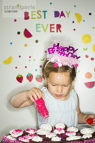 Caucasian girl sprinkling sprinkles on tray of cupcakes for birthday