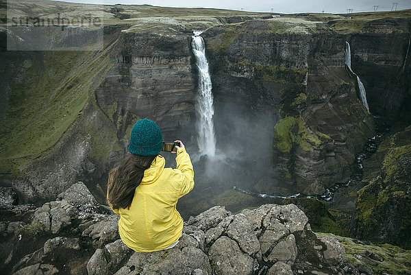 Kaukasische Frau sitzt auf einer Klippe und fotografiert einen Wasserfall