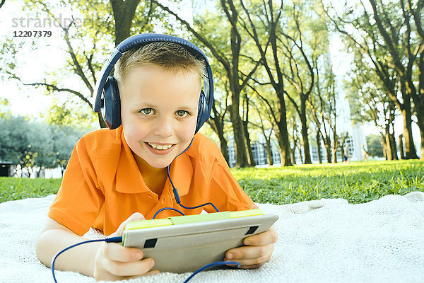 Smiling Caucasian boy laying on blanket in park listening to digital tablet