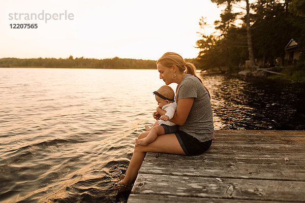 Mutter sitzt mit ihrer kleinen Tochter am Pier und schaut auf den See
