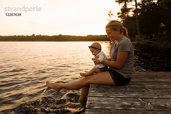 Mutter sitzt mit ihrer kleinen Tochter am Pier und planscht mit den Füßen im See