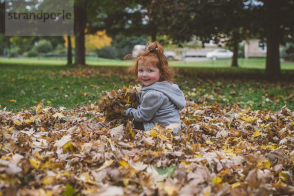 Porträt eines rothaarigen weiblichen Kleinkindes im Park mit Bündeln von Herbstblättern
