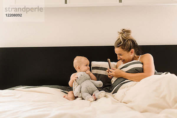 Mutter fotografiert Baby-Tochter im Bett sitzend mit Plüschtier