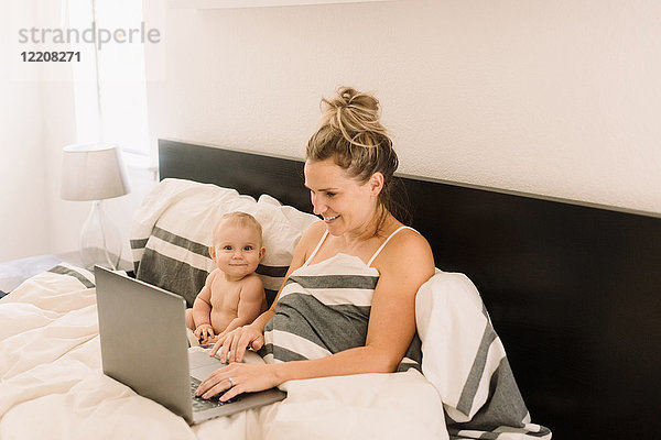 Porträt eines süßen Mädchens  das im Bett sitzt  während die Mutter einen Laptop benutzt