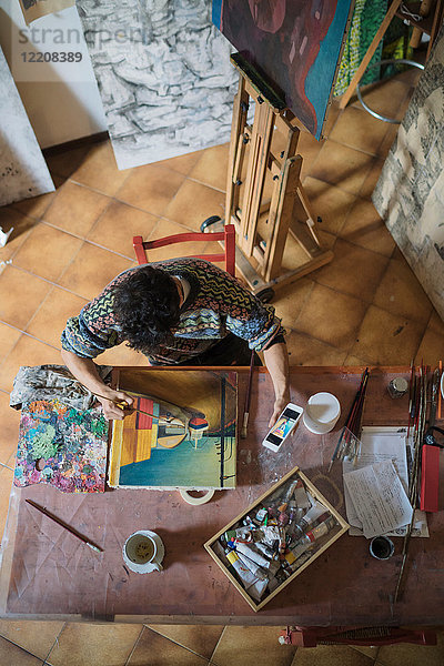 Männlicher Künstler schaut auf Smartphone  während er im Künstleratelier Leinwand malt  Draufsicht
