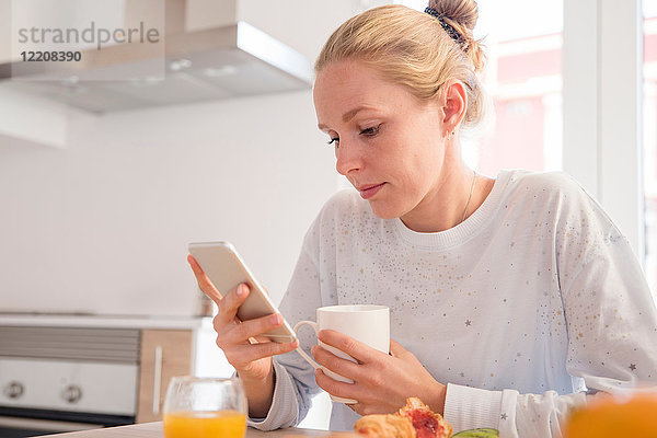 Junge Frau schaut auf Smartphone am Frühstückstisch